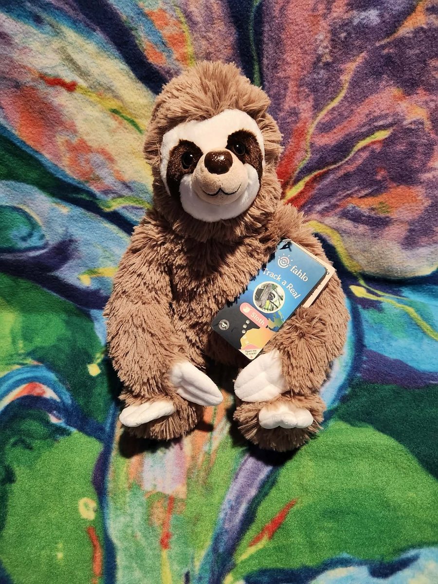 A plush toy sloth with a Fahlo tag reading Track a Real Sloth, resting on a colourful tie-dye blanket